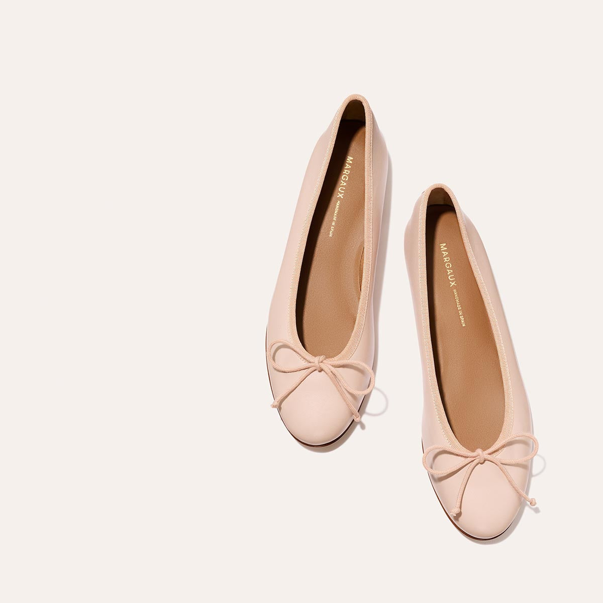 The Demi - Petal Nappa by Margaux are light pink ballet flats, handmade in Spain with bow details on the toes, displayed on a light neutral background.