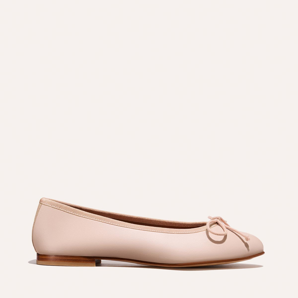 The Margaux Demi - Petal Nappa is a handmade nude ballet flat from Spain, featuring a petite bow on the toe, a low wooden heel, and displayed against a plain background.