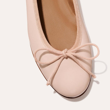 A close-up of The Demi - Petal Nappa by Margaux, a light pink ballet flat handmade in Spain with a rounded toe and matching bow, displayed on a simple, light background.