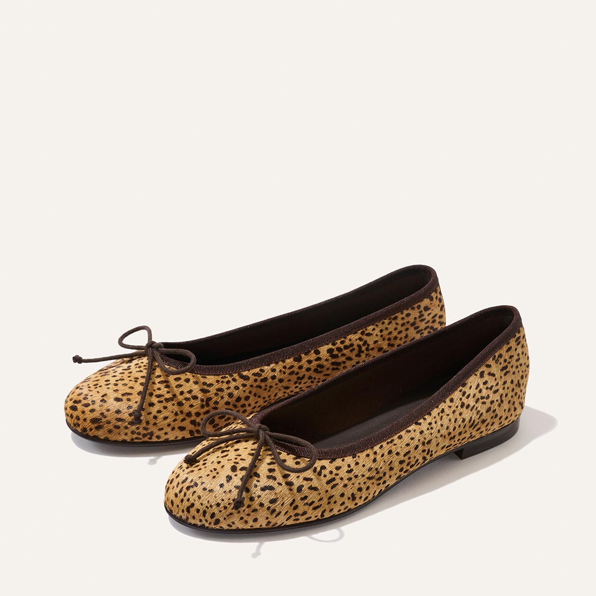 A pair of The Demi - Tan Mini Cheetah Haircalf ballet flats by Margaux, featuring a comfortable design with black trim and a black bow on the toe, handcrafted in Spain and shown against a plain white background.