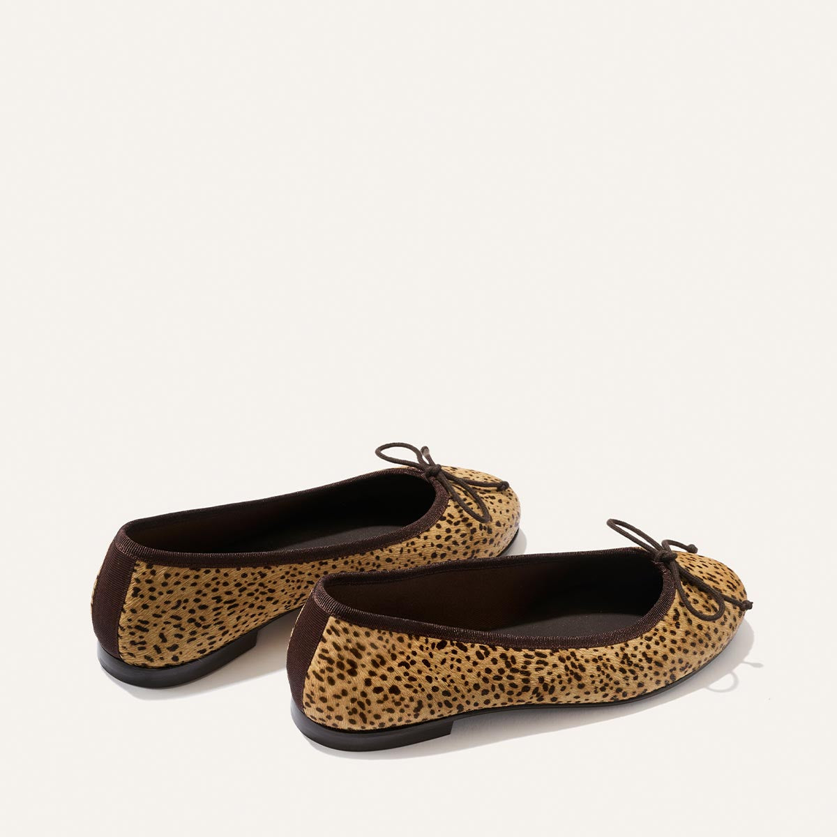Introducing The Demi - Tan Mini Cheetah Haircalf by Margaux: a pair of comfortable ballet flats featuring a chic mini cheetah print, black soles, brown trim, and delicate bows on the toes. Handmade in Spain and elegantly displayed against a plain white background.