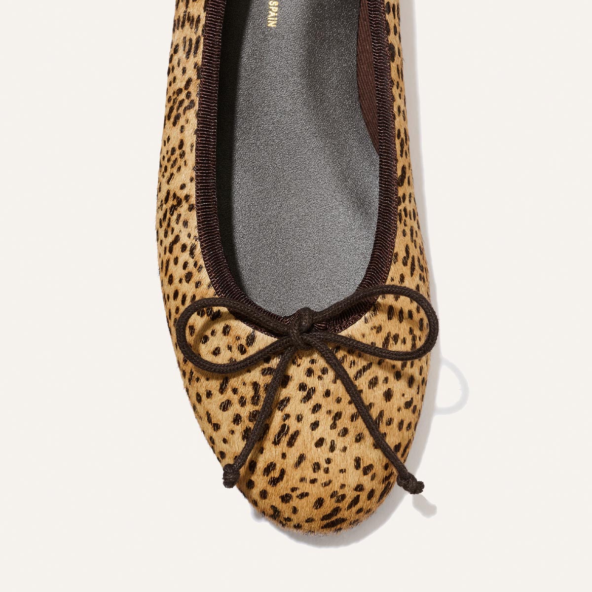A close-up of The Demi - Tan Mini Cheetah Haircalf by Margaux, a comfortable ballet flat handmade in Spain, featuring a chic black trim and a delicate black bow on the toe.