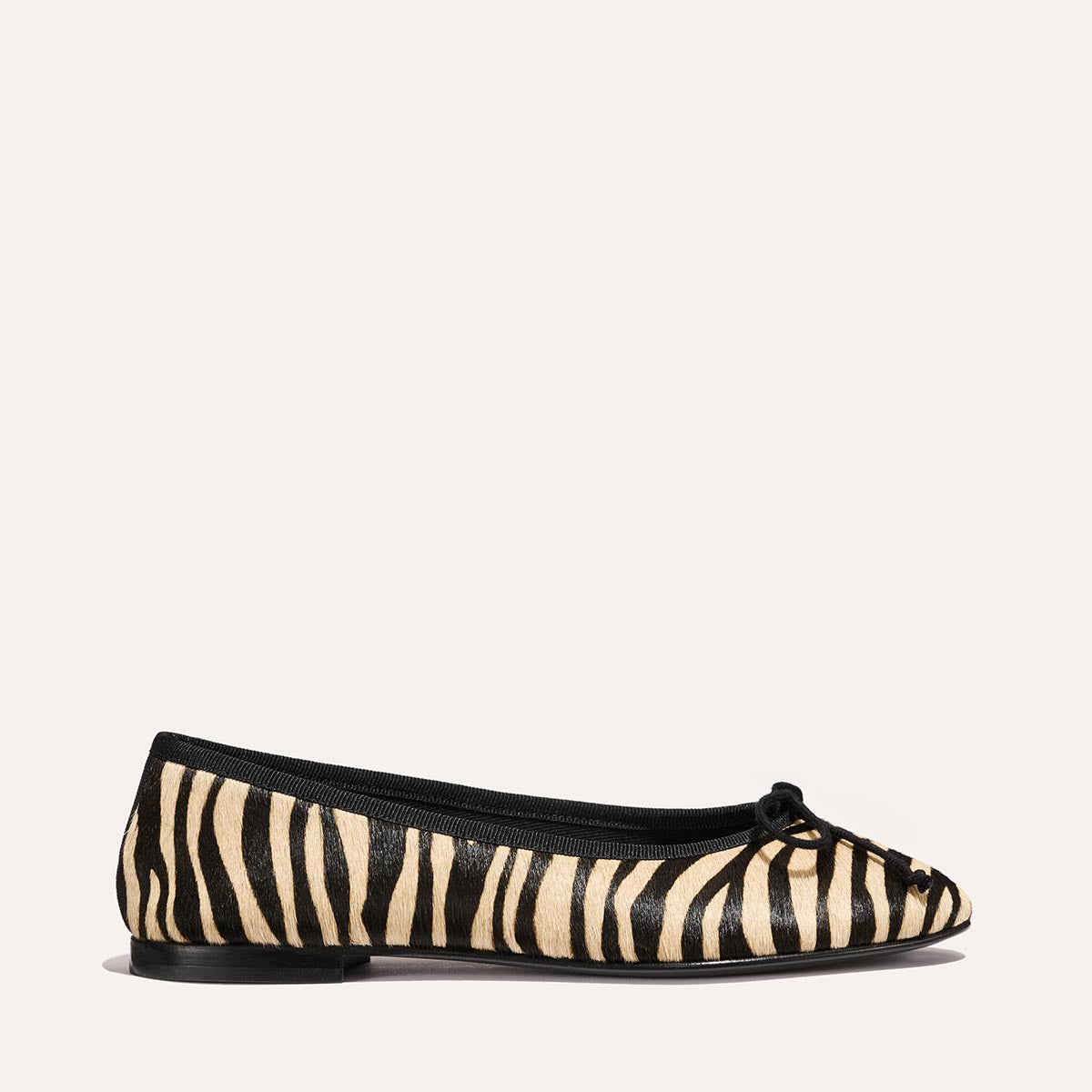 The Demi - Tan Zebra Haircalf by Margaux is a handmade ballet flat in Spain, featuring tan zebra haircalf, black trim and bow, and a flat black sole, displayed on a plain light background.