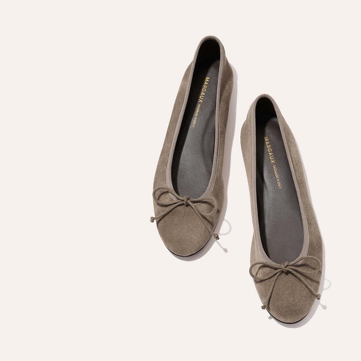 The Demi - Truffle Suede by Margaux: taupe suede ballet flats handmade in Spain, featuring rounded toes, decorative front bows, and black insoles—shown on a light background.