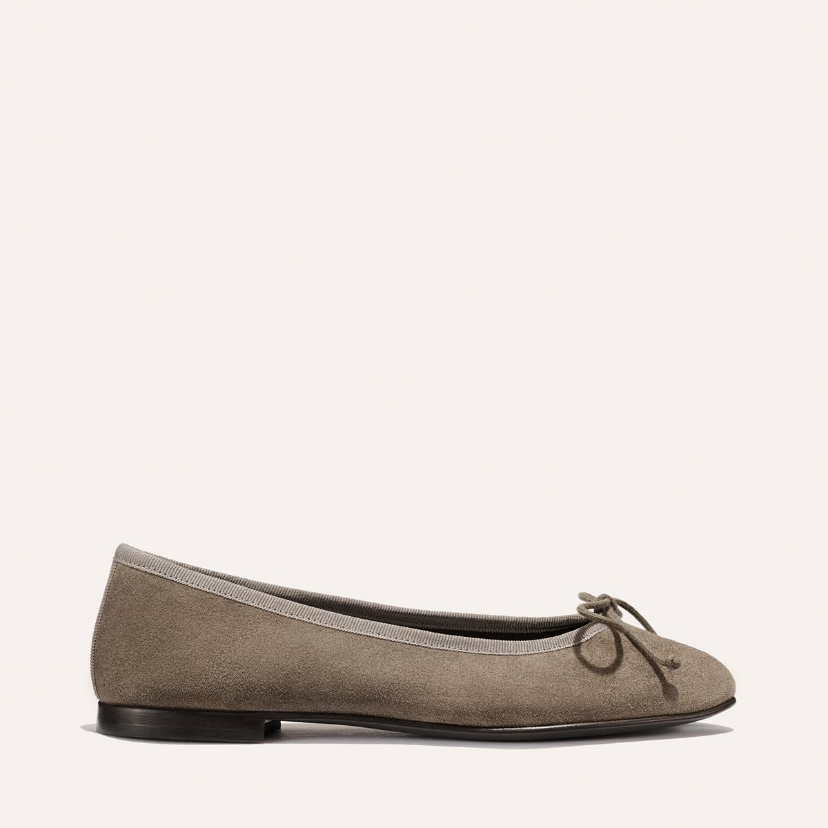 The Demi - Truffle Suede by Margaux is a comfortable taupe suede ballet flat, handmade in Spain with a rounded toe, front bow detail, and low black heel, shown against a plain light background.