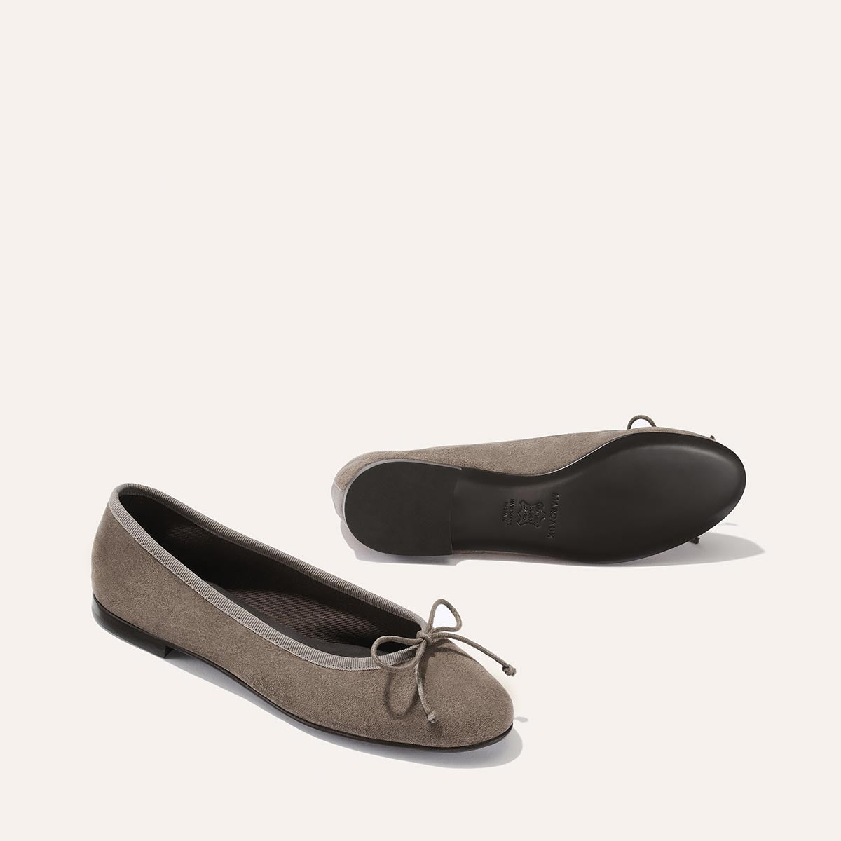 Two Truffle Suede “The Demi” ballet flats by Margaux are displayed against a plain light background—one standing upright and the other lying on its side to reveal the black sole. Both handmade in Spain, these taupe suede flats feature small bows on the front.