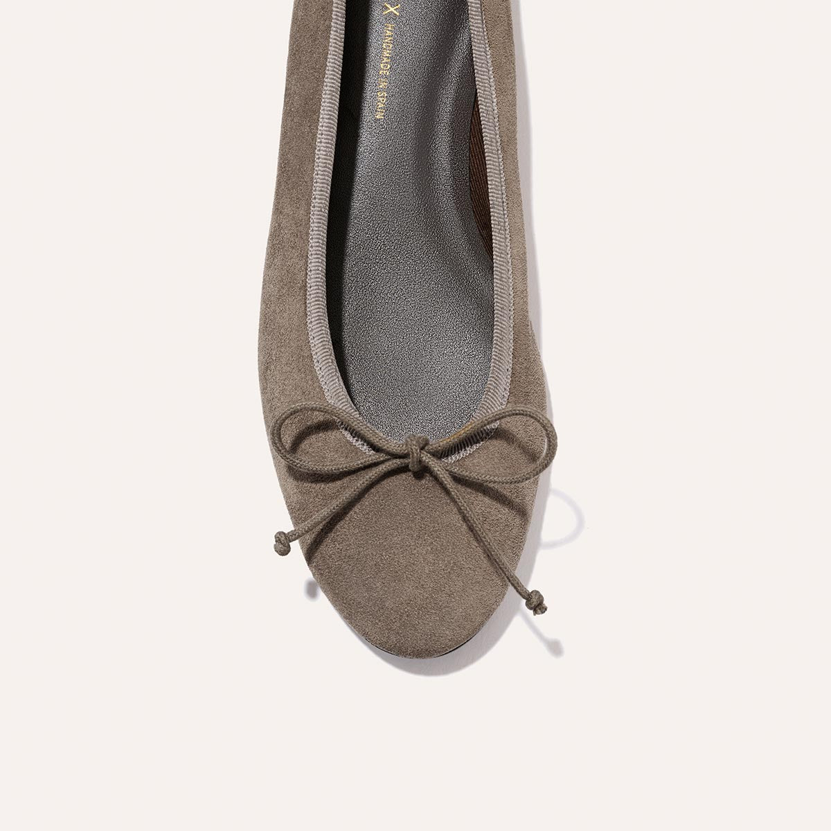A top view of Margaux’s The Demi – Truffle Suede ballet flat, handmade in Spain from taupe suede, featuring a matching bow at the toe and a gray insole, photographed on a light background.