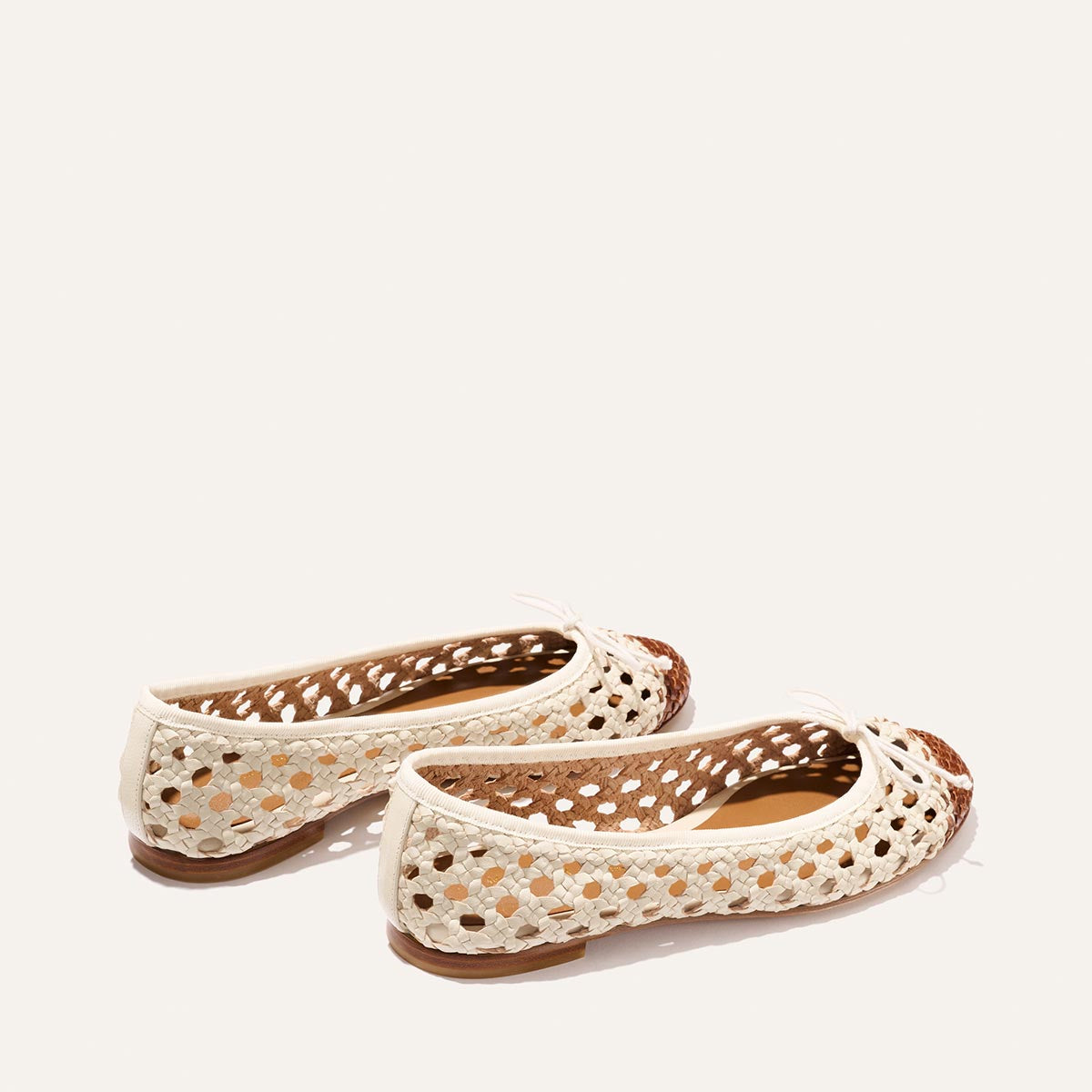 The Demi by Margaux in Ecru and Saddle features cream-colored woven leather ballet flats with small bows, perforated airy design, brown inner soles, and flat heels, displayed on a light background.