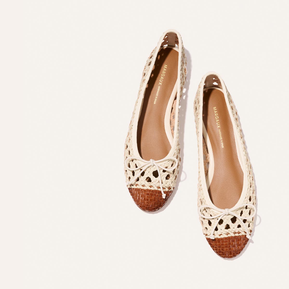 The Demi - Ecru and Saddle Woven Leather by Margaux are woven ballet flats with cream uppers, brown toes, and small bows on top, shown against a light background.
