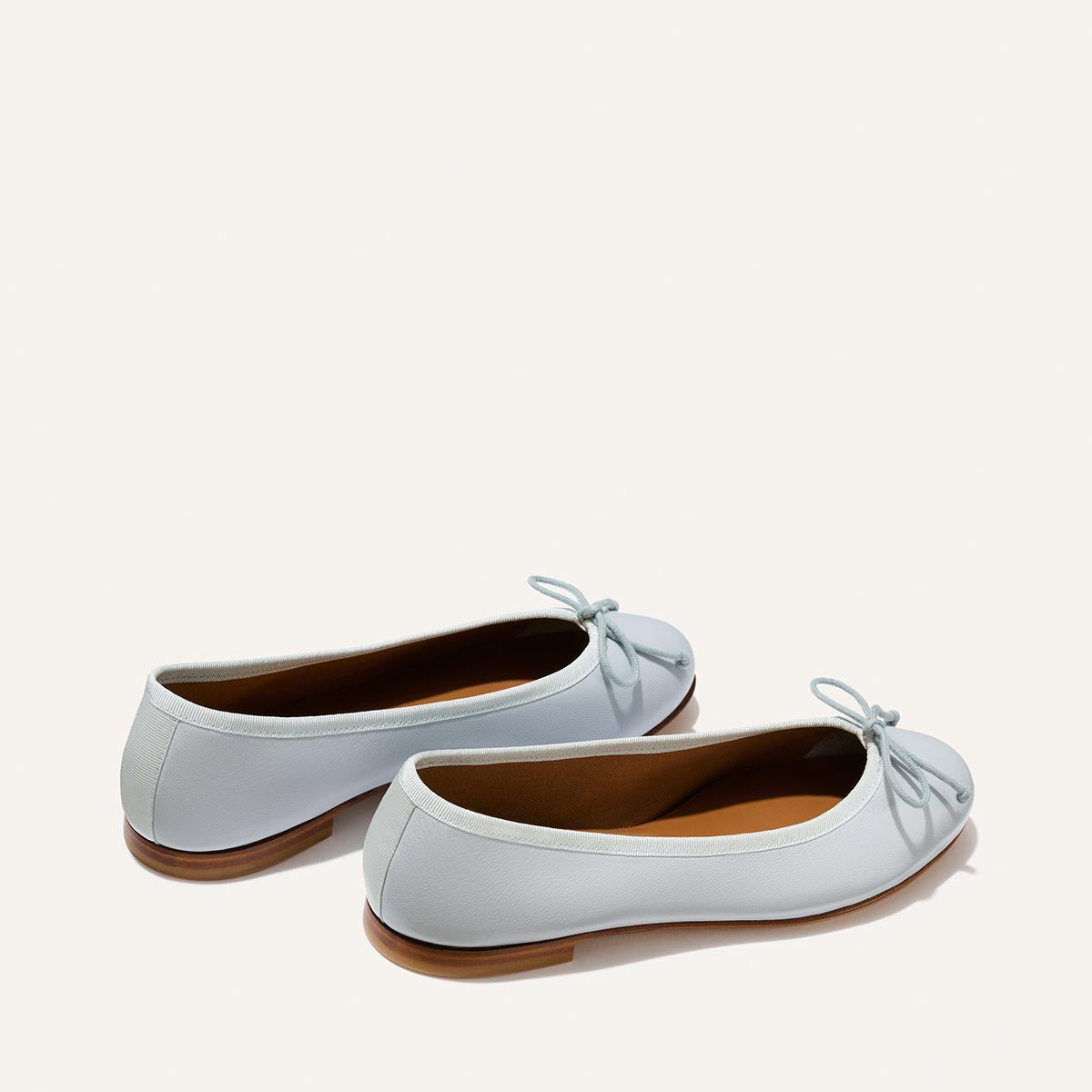 Margaux's The Demi - Mist Blue Nappa are comfortable ballet flats in light blue, handmade in Spain. They feature small bows on the toes, brown soles, and a soft, smooth texture, displayed on an off-white background.