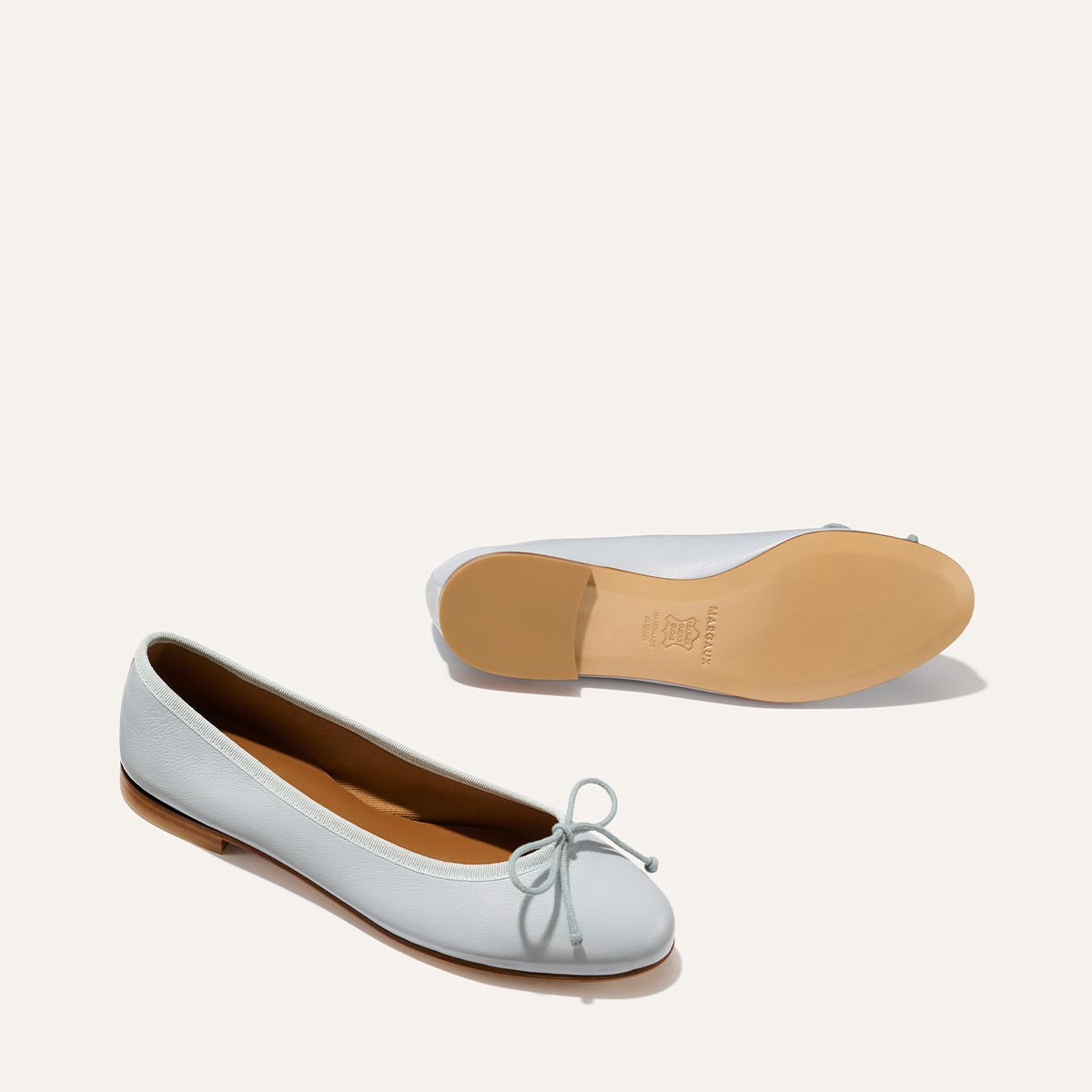 A pair of Margaux's The Demi - Mist Blue Nappa ballet flats, handmade in Spain, feature a small bow and tan sole; one shoe stands upright while the other displays its sole on a plain white background.