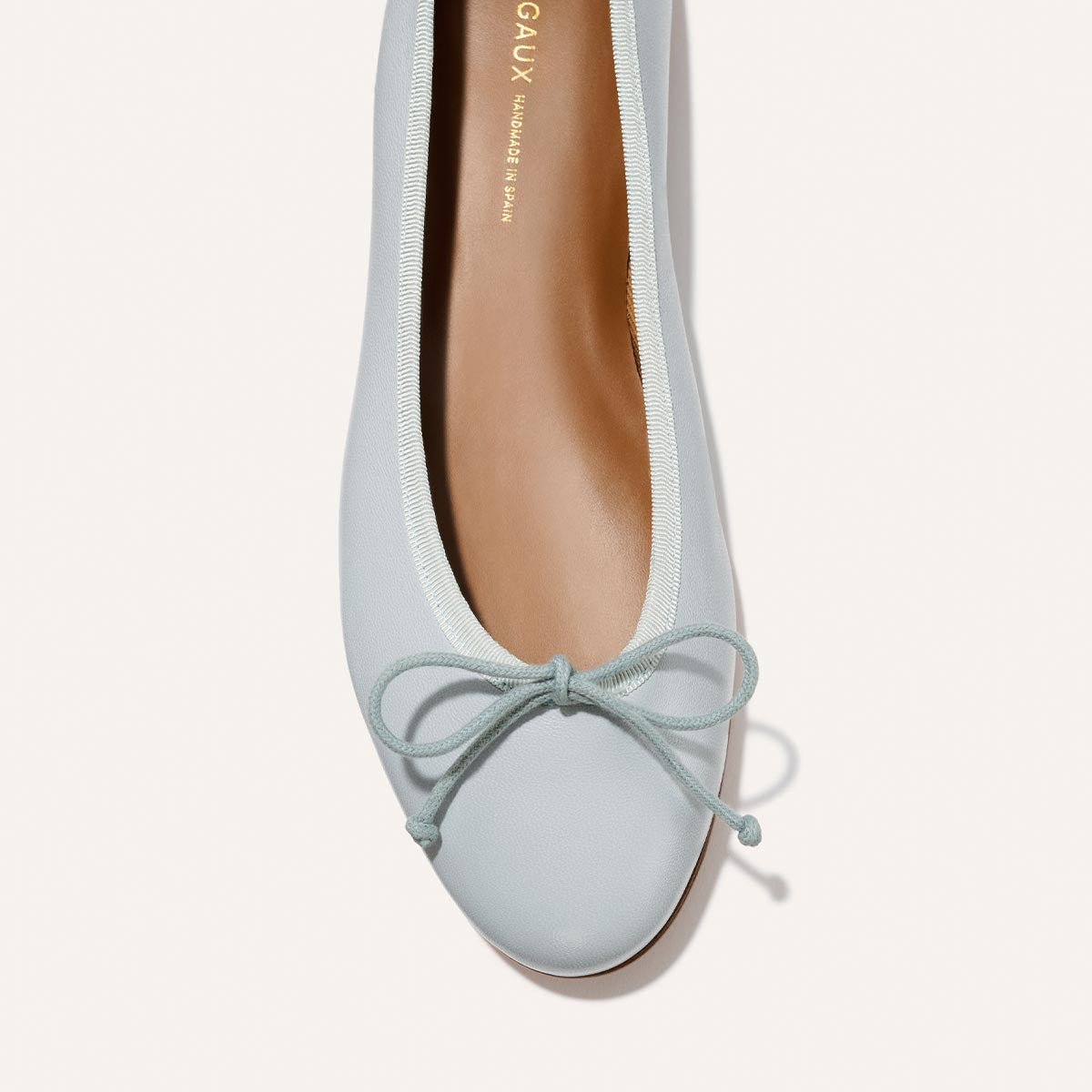 A close-up of The Demi - Mist Blue Nappa by Margaux: a comfortable, light blue ballet flat with a matching bow on the rounded toe, tan insole, and gold brand lettering inside. Handmade in Spain.