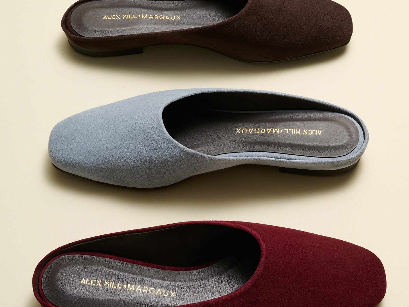 Three pairs of slippers in brown, blue, and red with 'Alex Mill & Margaux' branding on a beige background.