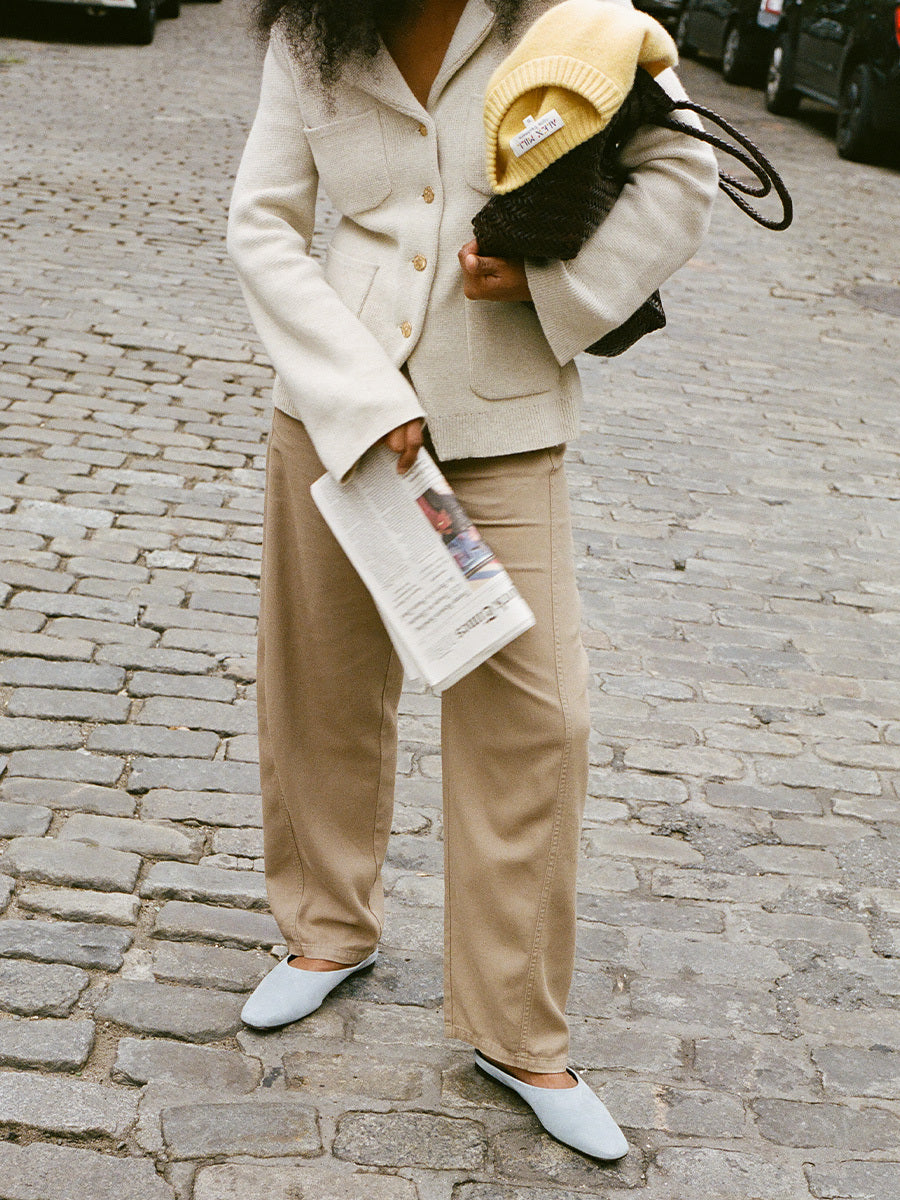 Person wearing a beige blazer and pants, holding a newspaper and a yellow beanie on a cobblestone street.