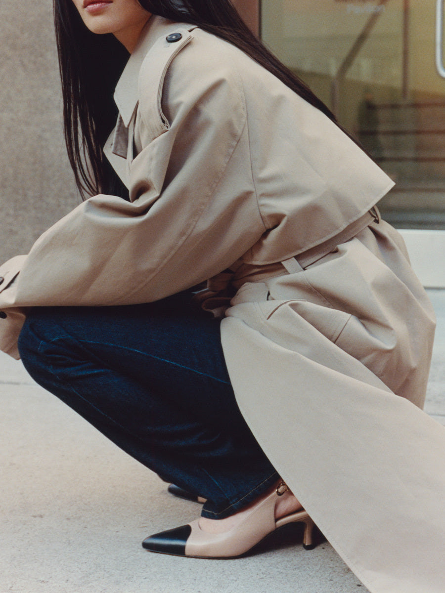 Person wearing a beige trench coat and dark jeans, squatting on a concrete surface.