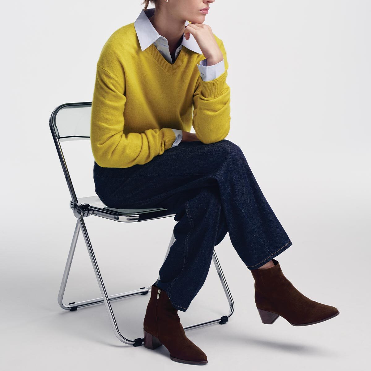 A person in a yellow sweater, collared shirt, and dark trousers sits on a transparent folding chair wearing Chocolate Suede The Downtown Boot with a pointed toe and a stacked leather heel.