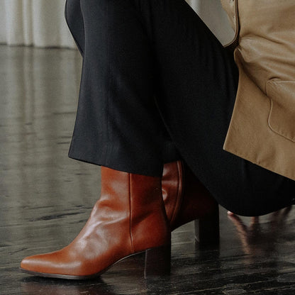 A person in dark pants and a tan ankle-length coat sits on a chair, wearing Margaux’s The Downtown Boot - Saddle Calf; the chic brown leather heeled boots complement the dark wood floor and white curtain backdrop.