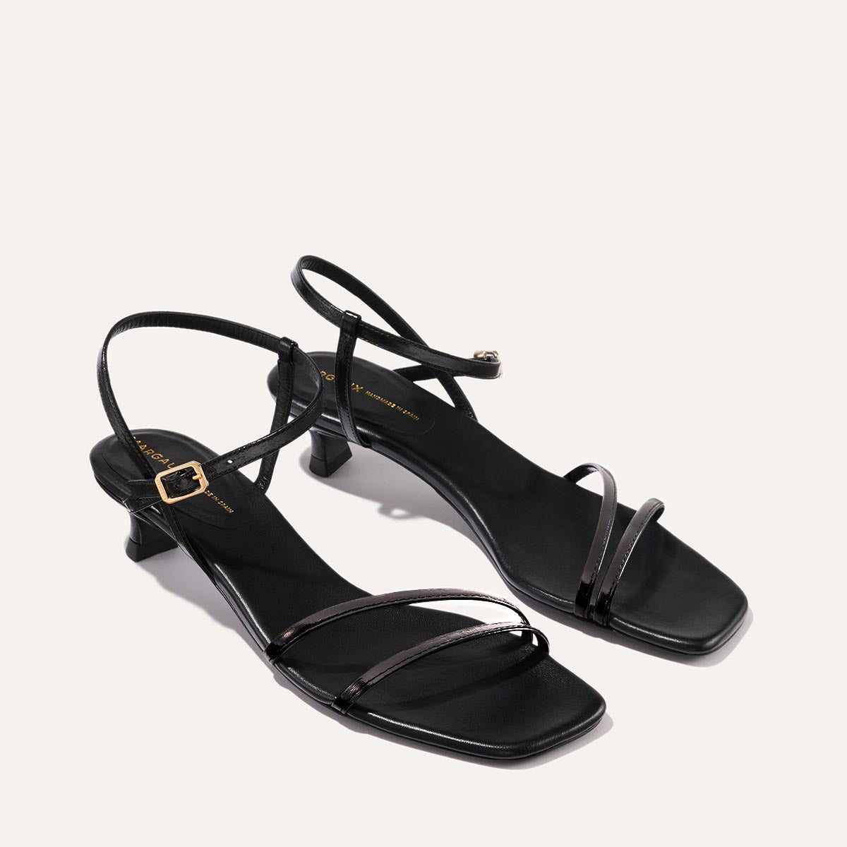 The Dylan Sandal in shiny Black Crinkle-Gloss Nappa, featuring asymmetrical straps around the ankle and toes and a walkable 1-inch stiletto heel.