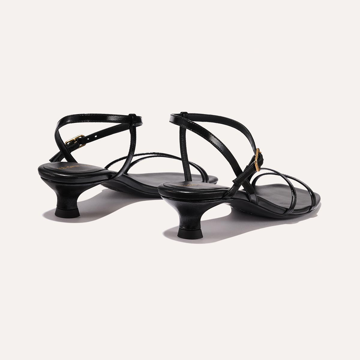 The Dylan Sandal in shiny Black Crinkle-Gloss Nappa, featuring asymmetrical straps around the ankle and toes and a walkable 1-inch stiletto heel.