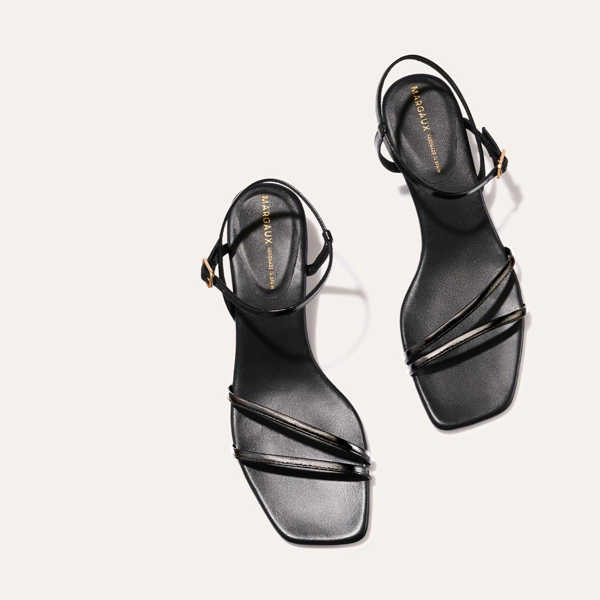 The Dylan Sandal in shiny Black Crinkle-Gloss Nappa, featuring asymmetrical straps around the ankle and toes and a walkable 1-inch stiletto heel.