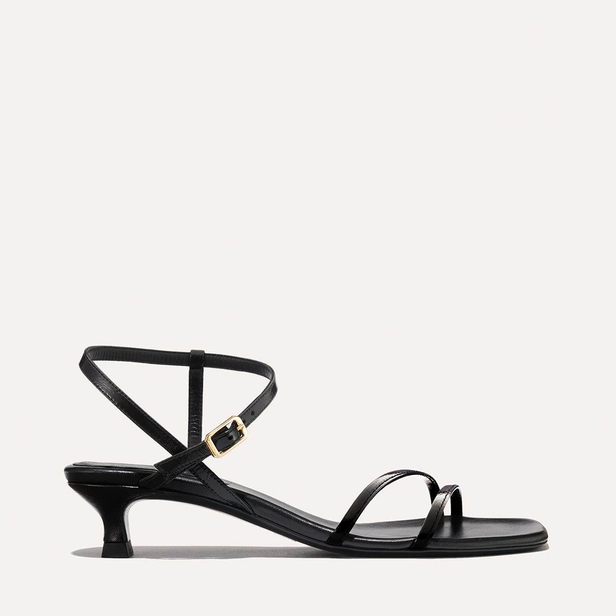 The Dylan Sandal in shiny Black Crinkle-Gloss Nappa, featuring asymmetrical straps around the ankle and toes and a walkable 1-inch stiletto heel.