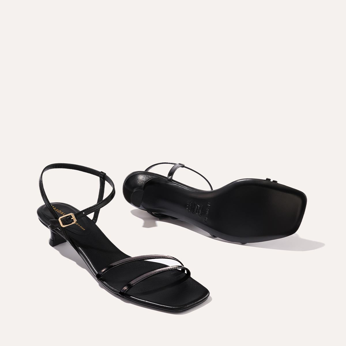 The Dylan Sandal in shiny Black Crinkle-Gloss Nappa, featuring asymmetrical straps around the ankle and toes and a walkable 1-inch stiletto heel.