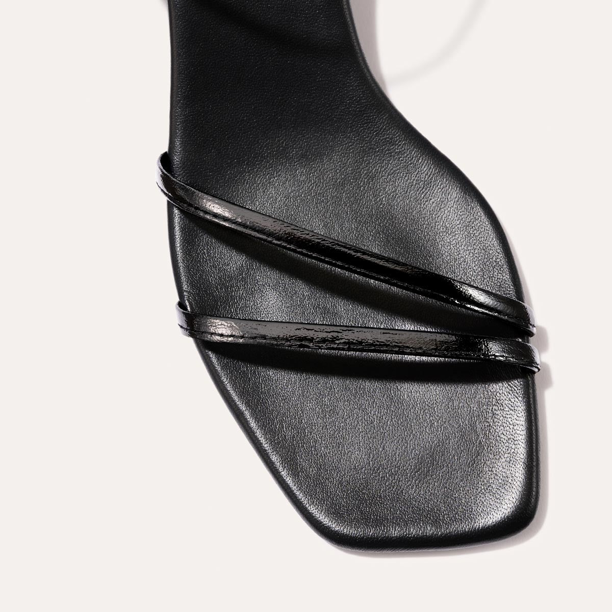 The Dylan Sandal in shiny Black Crinkle-Gloss Nappa, featuring asymmetrical straps around the ankle and toes and a walkable 1-inch stiletto heel.