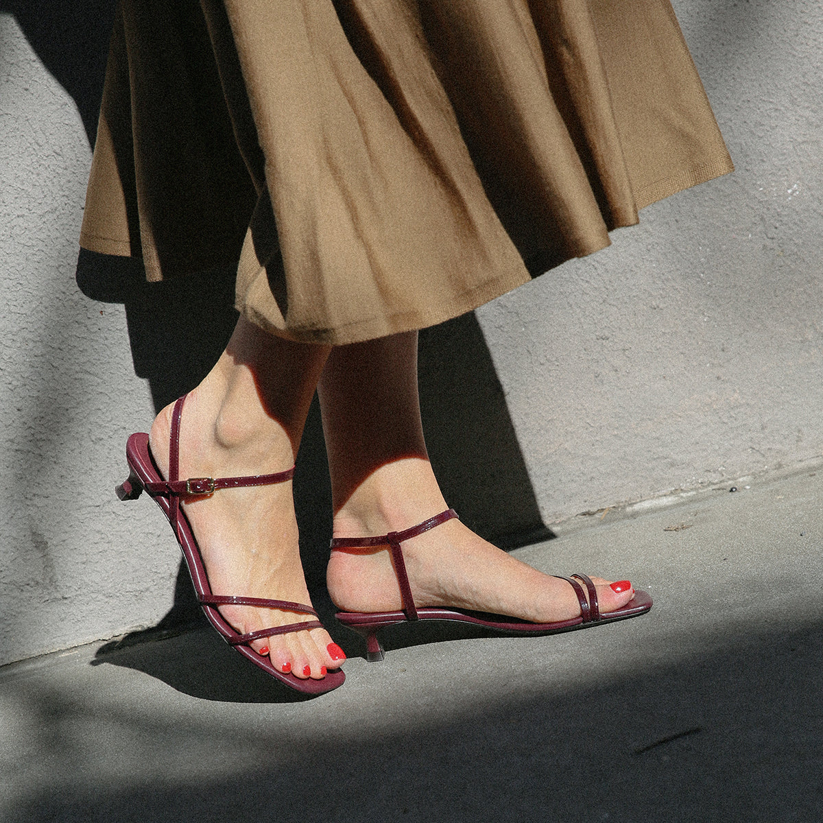 The Dylan Sandal in shiny Burgundy Patent, featuring asymmetrical straps around the ankle and toes and a walkable 1-inch stiletto heel.