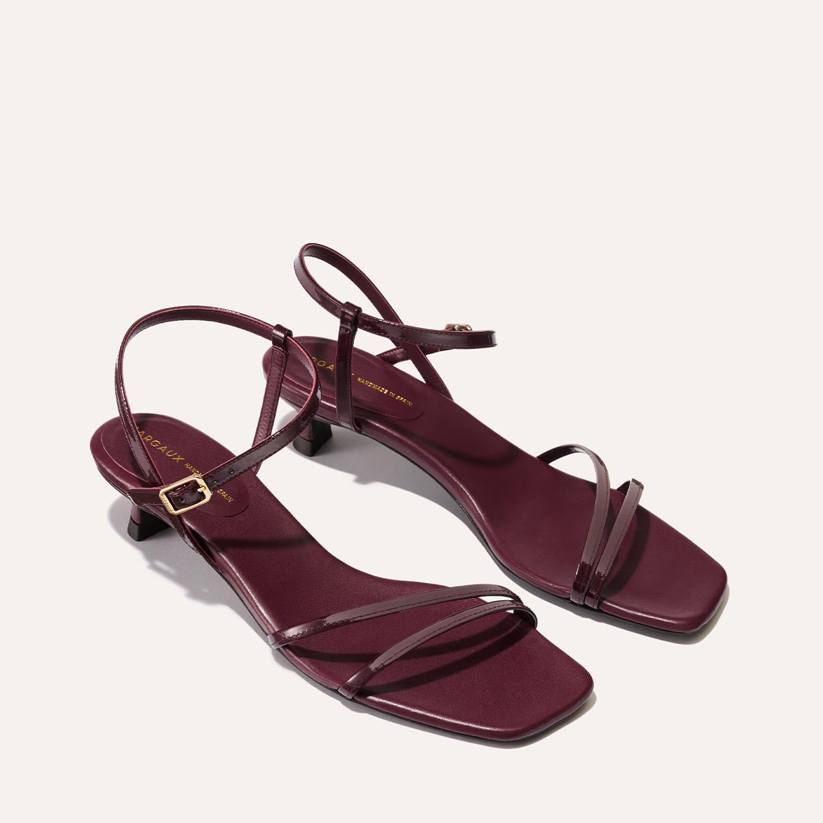 The Dylan Sandal in shiny Burgundy Patent, featuring asymmetrical straps around the ankle and toes and a walkable 1-inch stiletto heel.