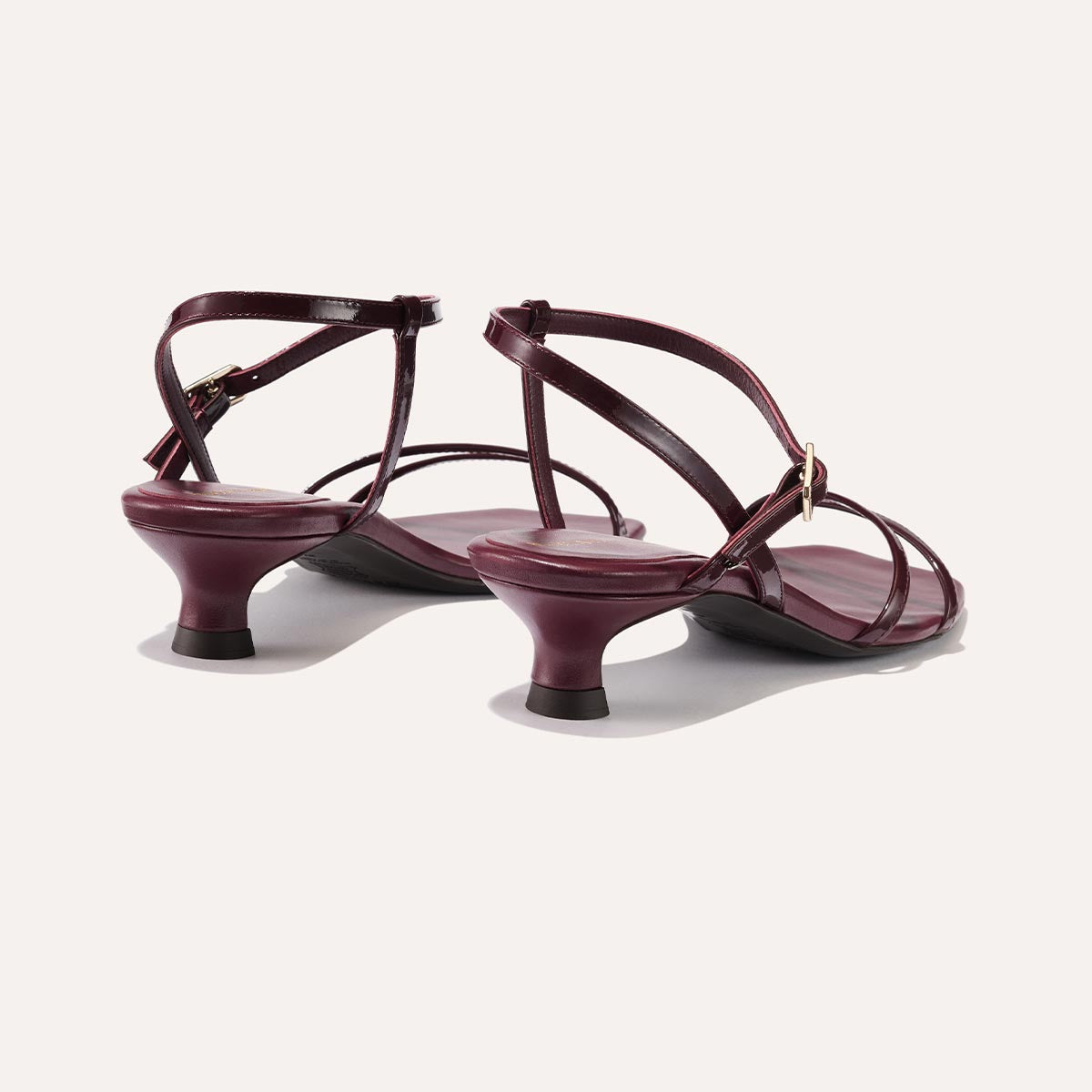 The Dylan Sandal in shiny Burgundy Patent, featuring asymmetrical straps around the ankle and toes and a walkable 1-inch stiletto heel.