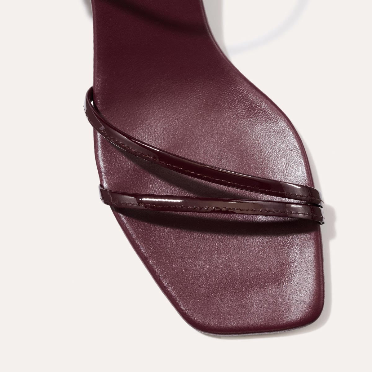 The Dylan Sandal in shiny Burgundy Patent, featuring asymmetrical straps around the ankle and toes and a walkable 1-inch stiletto heel.