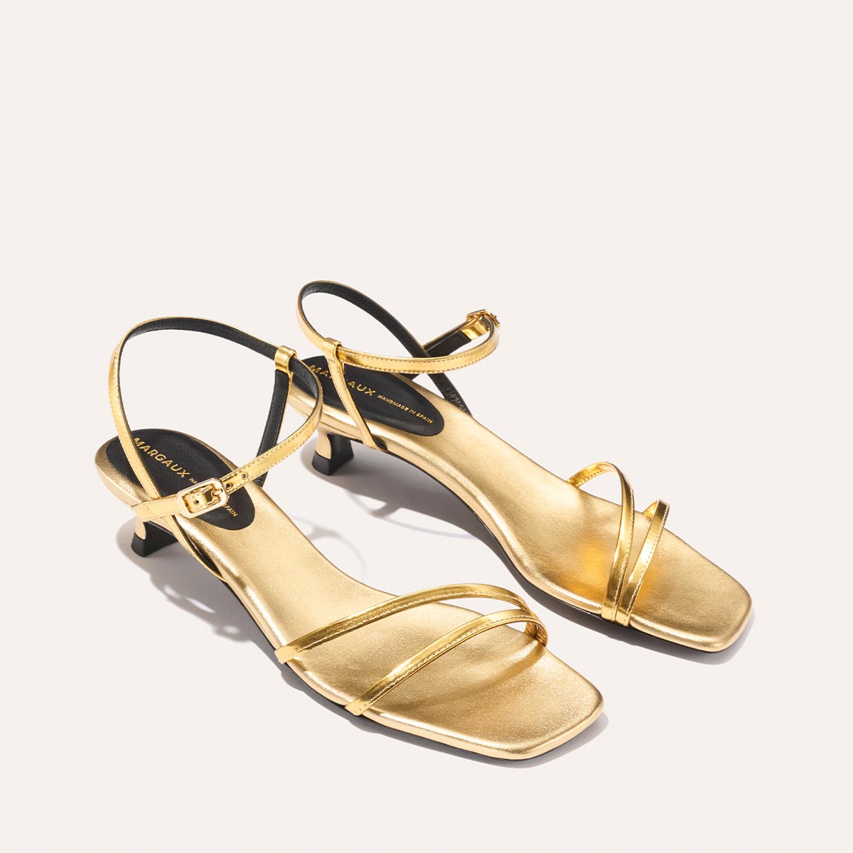 The Dylan Sandal - Gold Mirror by margaux-development: gold strappy heeled sandals with square toes, small heels, ankle straps, and a minimalist design shown on a light background.