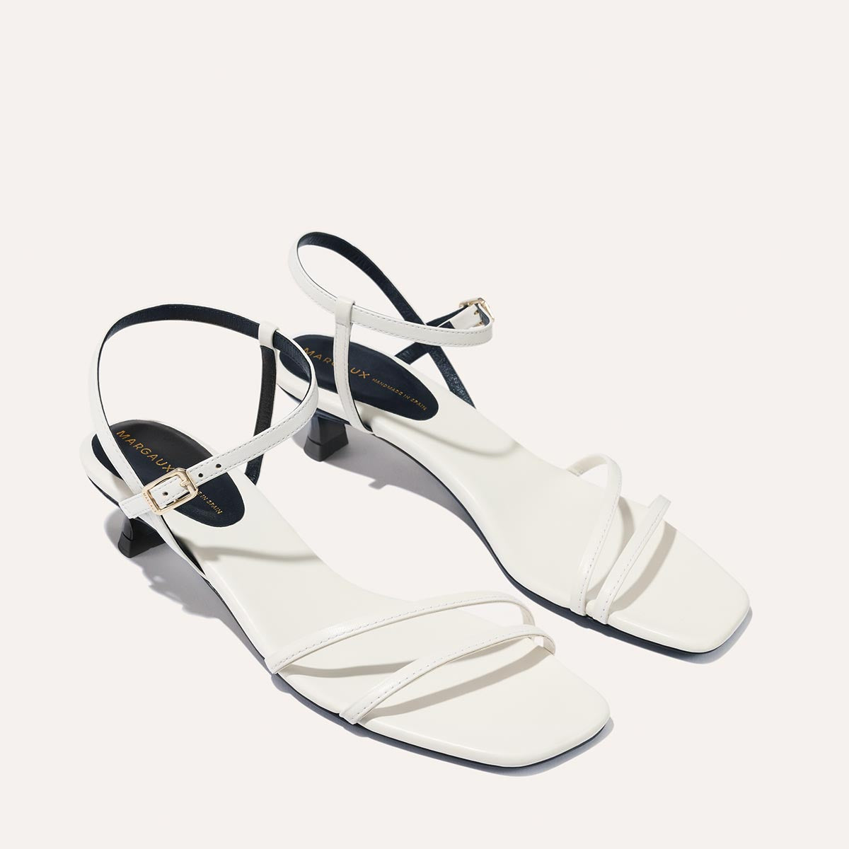 The Dylan Sandal in Ivory Nappa, featuring asymmetrical straps around the ankle and toes and a walkable 1-inch stiletto heel.