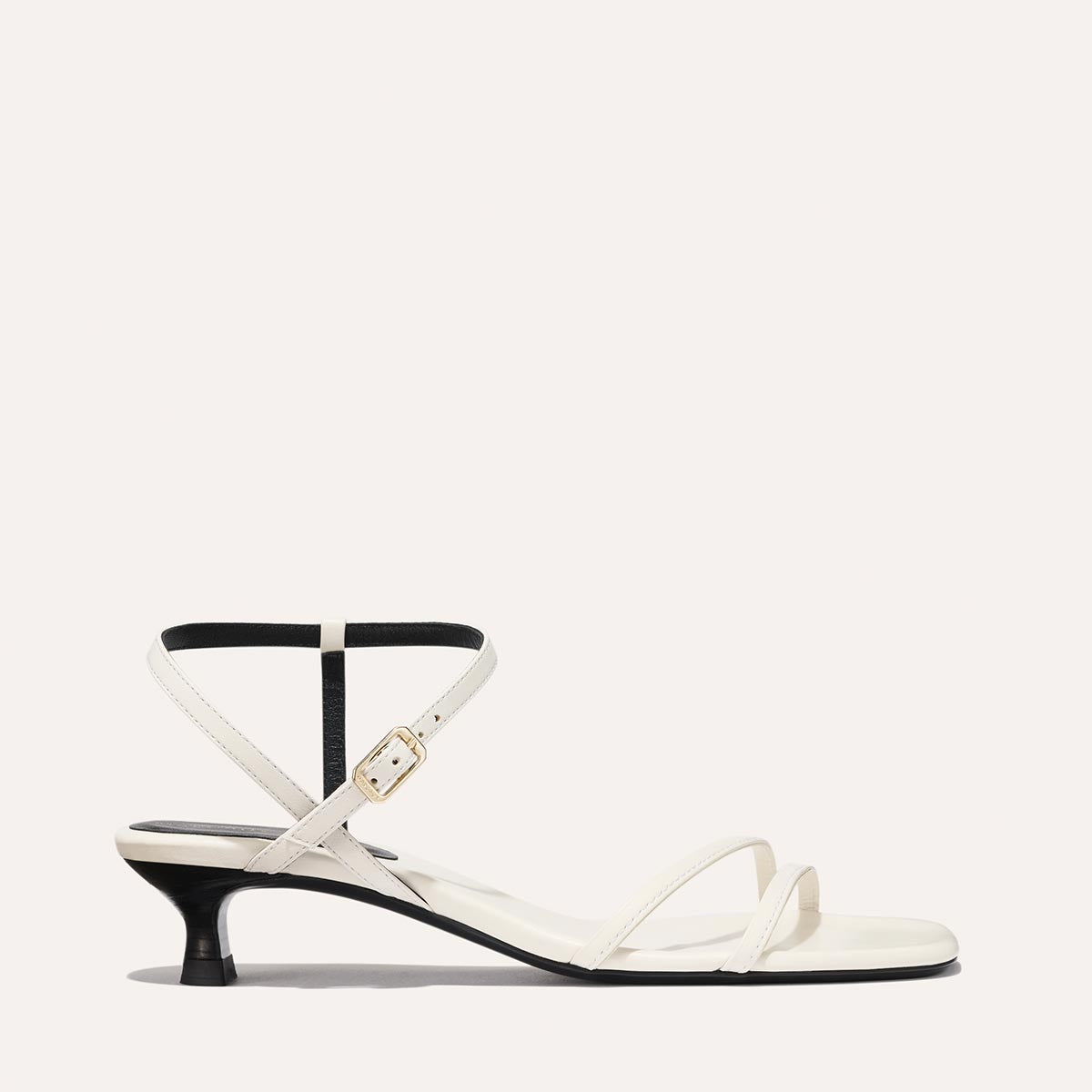 The Dylan Sandal in Ivory Nappa, featuring asymmetrical straps around the ankle and toes and a walkable 1-inch stiletto heel.