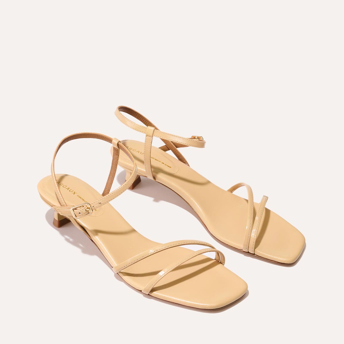 The Dylan Sandal in shiny Sand Crinkle-Gloss Nappa, featuring asymmetrical straps around the ankle and toes and a walkable 1-inch stiletto heel.