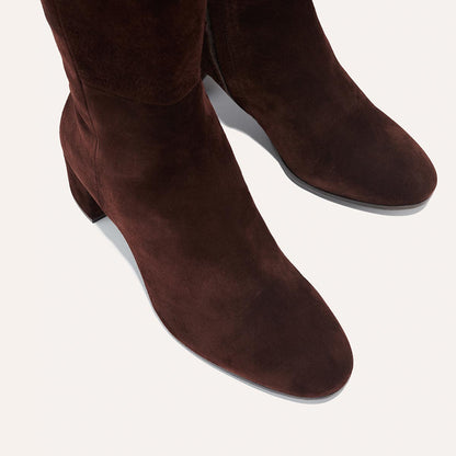 The Edie Boot in Chocolate Suede, a sleek knee-high silhouette complemented by a comfortable 2.5-inch block heel and a hidden interior zipper for a fitted look.