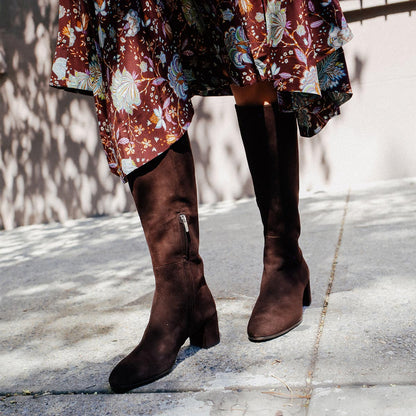 The Edie Boot in Chocolate Suede, a sleek knee-high silhouette complemented by a comfortable 2.5-inch block heel and a hidden interior zipper for a fitted look.
