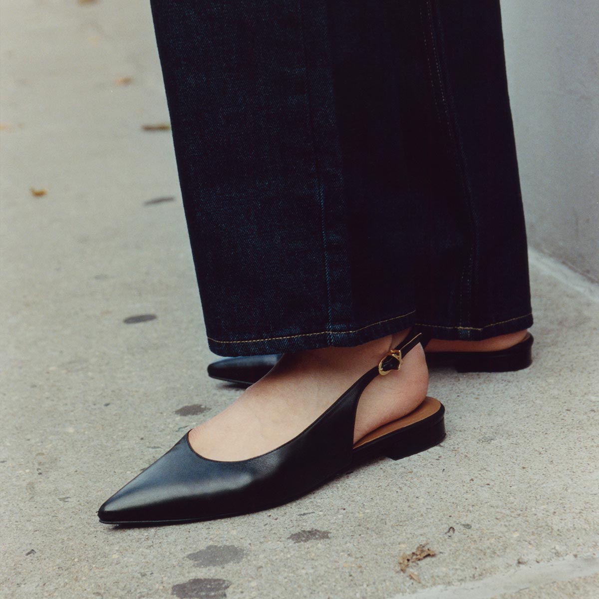 A person wearing dark jeans and The Elise - Black Nappa slingback flats by margaux-development, featuring a cap toe and leather sole, stands on a concrete sidewalk.