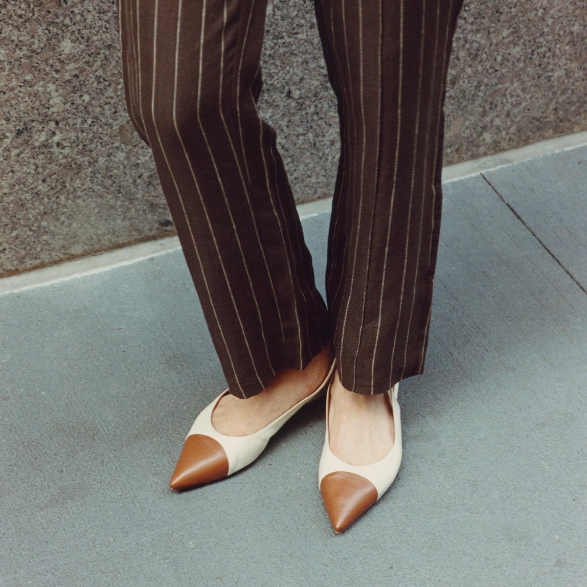 A person stands on a gray sidewalk by a rough stone wall, wearing brown pinstriped pants and margaux-development’s The Elise flats in cotton and Saddle Nappa.