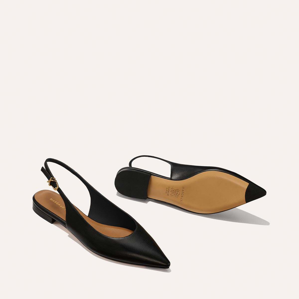 The Elise - Black Nappa by margaux-development: a pair of black pointed-toe slingback flats with low heels and tan soles, displayed on a light background—one upright, the other on its side showing the sole.
