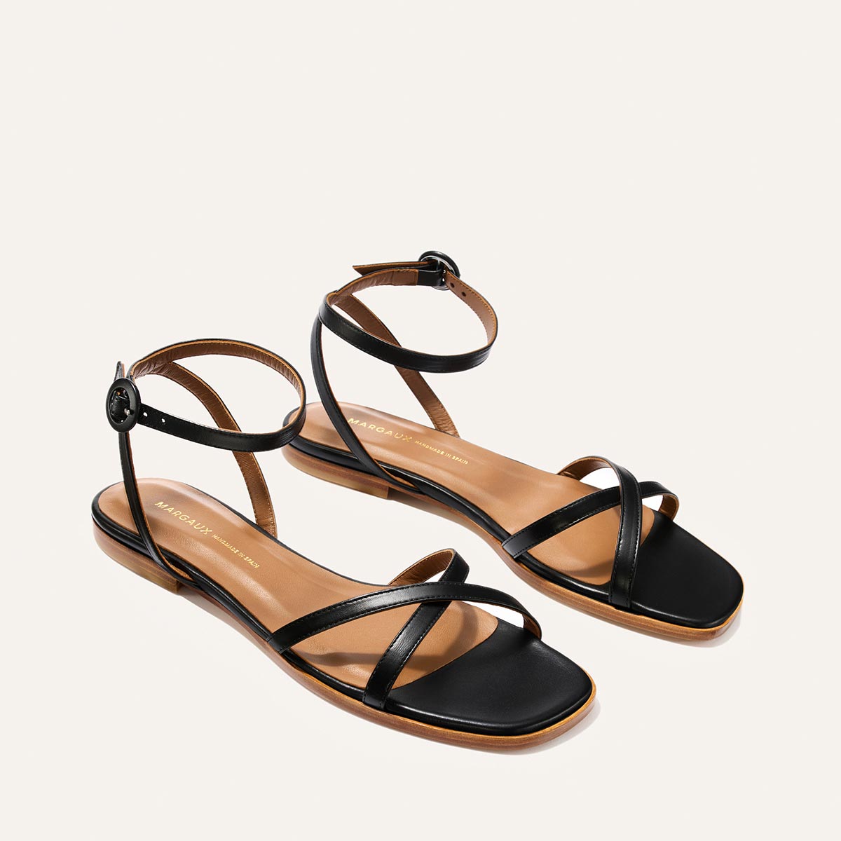 The Flat Sandal - Black Nappa by Margaux features thin crisscross and ankle buckle straps, plus brown foam-cushioned insoles, shown on a light background.