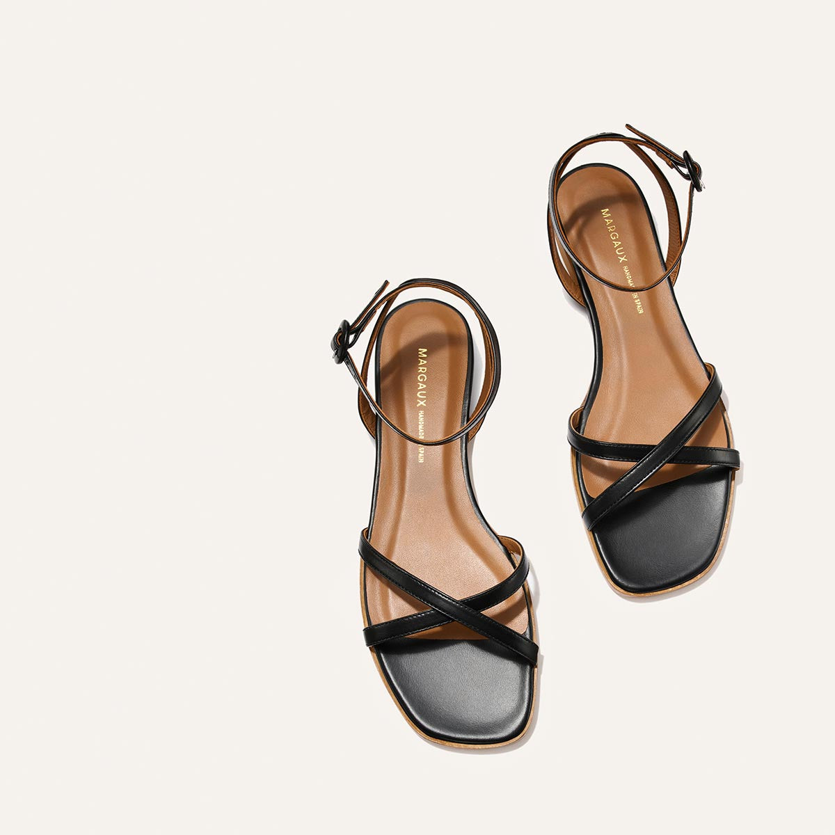 The Flat Sandal - Black Nappa by Margaux: minimalist black barely-there sandals with ankle straps, square toes, and brown insoles displayed on a light background—a timeless style for any occasion.