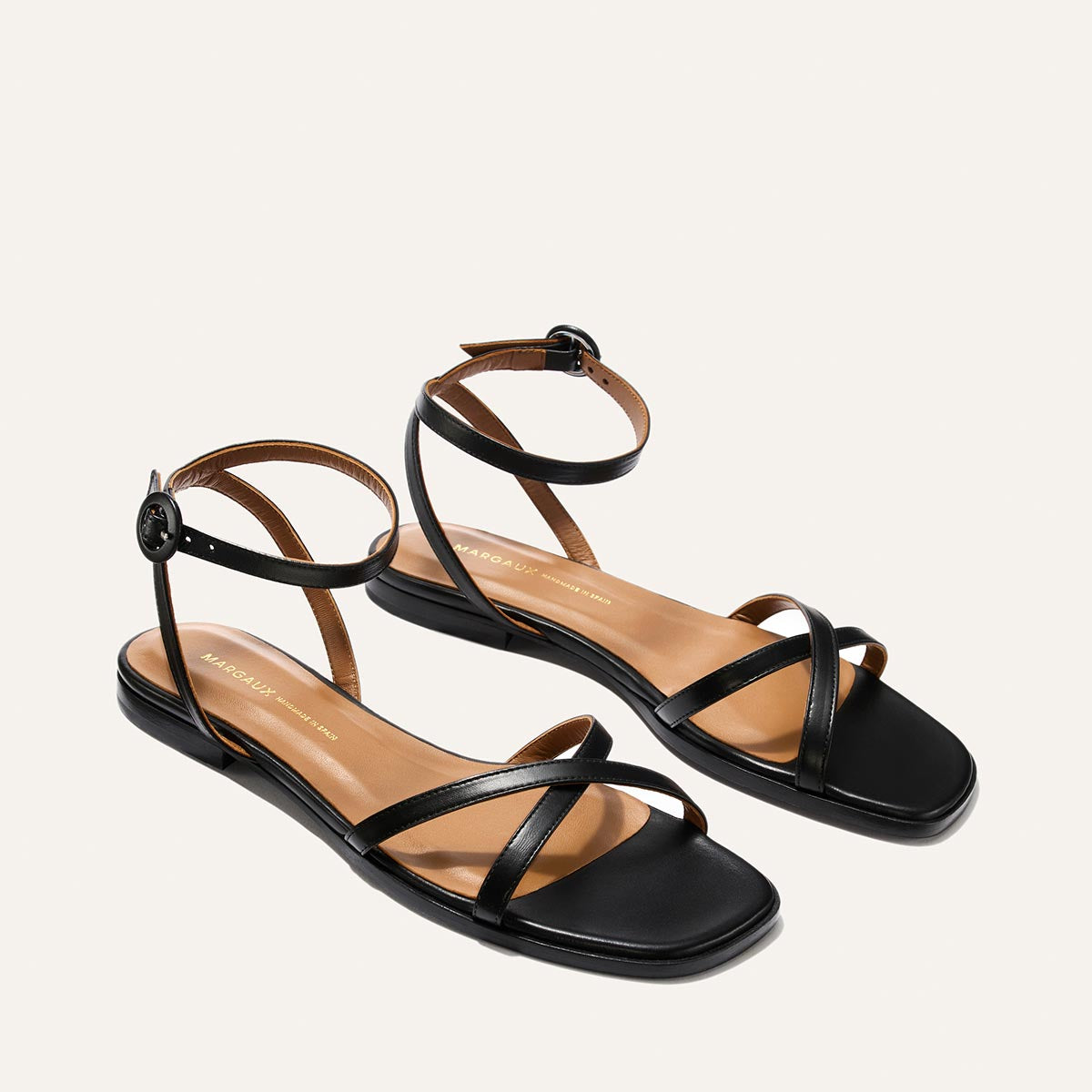 The Flat Sandal - Black Nappa by Margaux: minimalist black sandals with ankle straps, small buckles, and a tan foam-cushioned insole, shown on a plain light background.