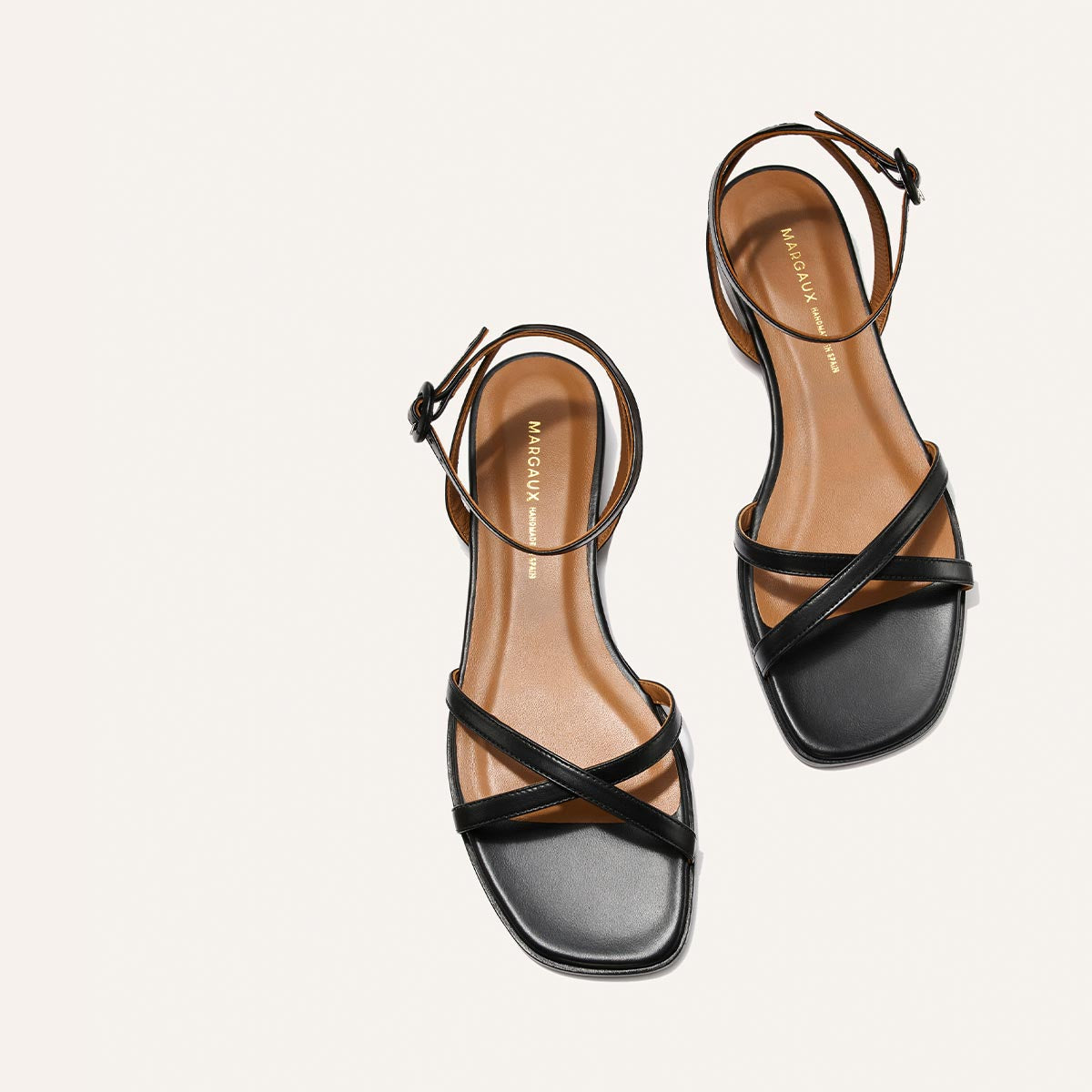A pair of black flat sandals, The Flat Sandal - Black Nappa by Margaux, with tan foam-cushioned insoles, ankle straps, and square toes are displayed on a white background. "Margaux" is visible on the insoles.