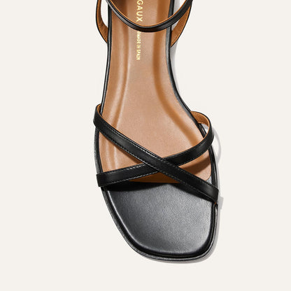 A close-up of The Flat Sandal - Black Nappa by Margaux, featuring thin crisscross black leather straps, a square toe, and a brown foam-cushioned insole with the Margaux logo in gold for a timeless touch.