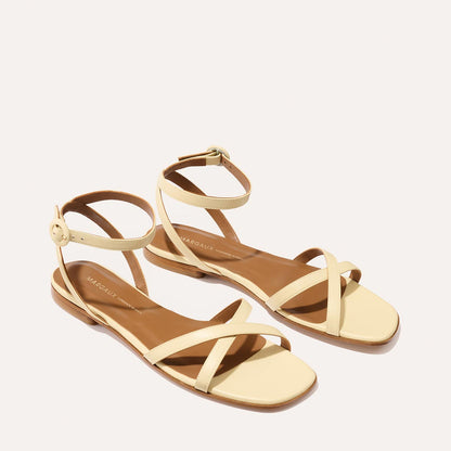 Margaux’s The Flat Sandal - Butter Nappa features beige strappy flats with ankle straps, buckle closure, brown insoles for comfort, and thin crisscrossing toe and foot straps.
