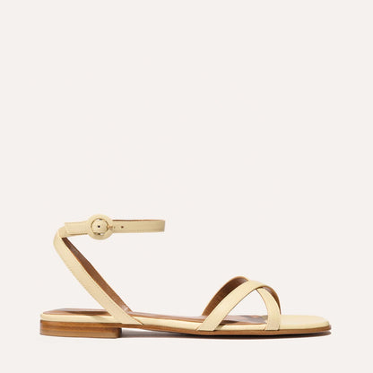 The Flat Sandal - Butter Nappa by Margaux is a cream-hued sandal with comfortable diagonal straps, a slim ankle strap with side buckle, and a light brown sole with low heel. Displayed on a plain, light background.