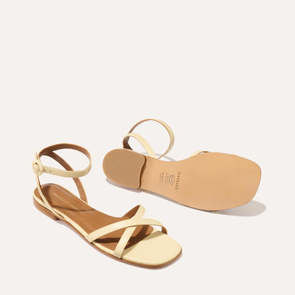 A pair of Margauxs The Flat Sandal in Butter Nappa, featuring light beige strappy flats with ankle straps—one upright to show the top design and the other flipped to reveal the sole against a plain light background.