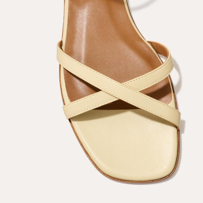 A close-up of Margauxs The Flat Sandal - Butter Nappa, featuring cream crisscross straps and a square toe on a tan comfort footbed, set against a light background.