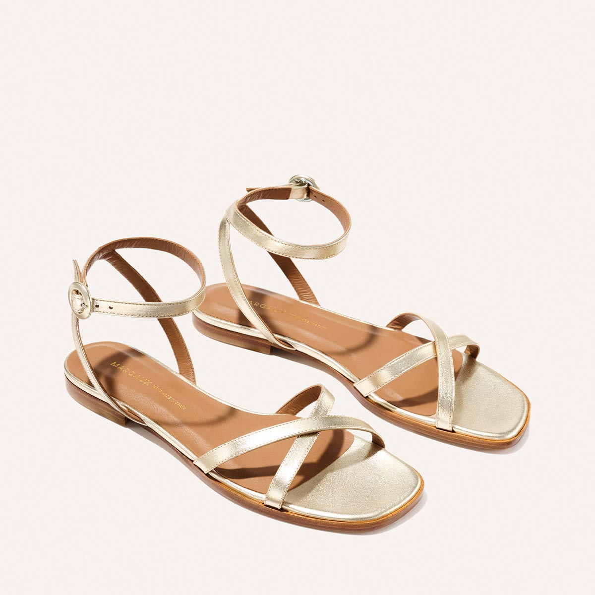 The Flat Sandal - Champagne by Margaux features metallic gold straps, foam-cushioned insoles, ankle buckle closures, and tan soles, shown on a light background.